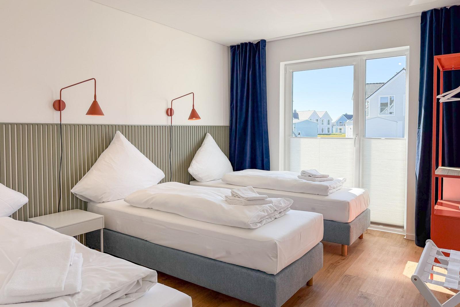 Zwei Betten mit weißen Bettwäschen, Fenster mit Blick auf Häuser, blaue Vorhänge und rote Wandlampen.