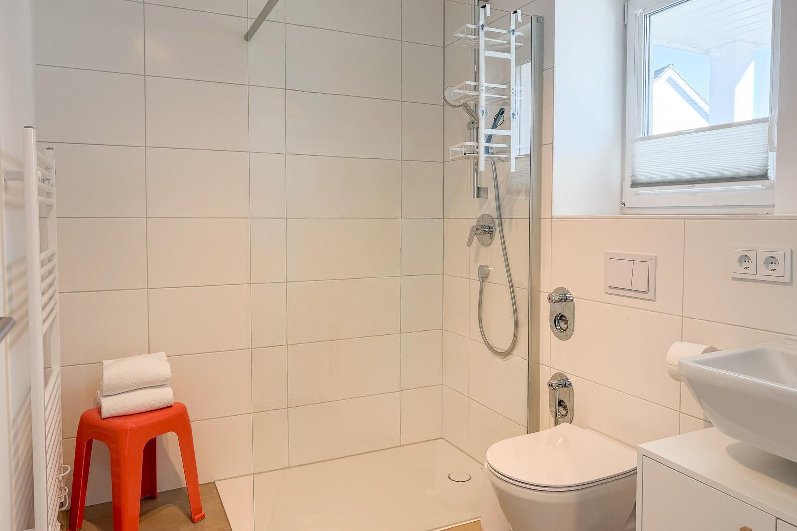 Dusche mit Duschbrause, Toilette und Waschbecken. Ein roter Hocker steht neben dem Heizkörper.