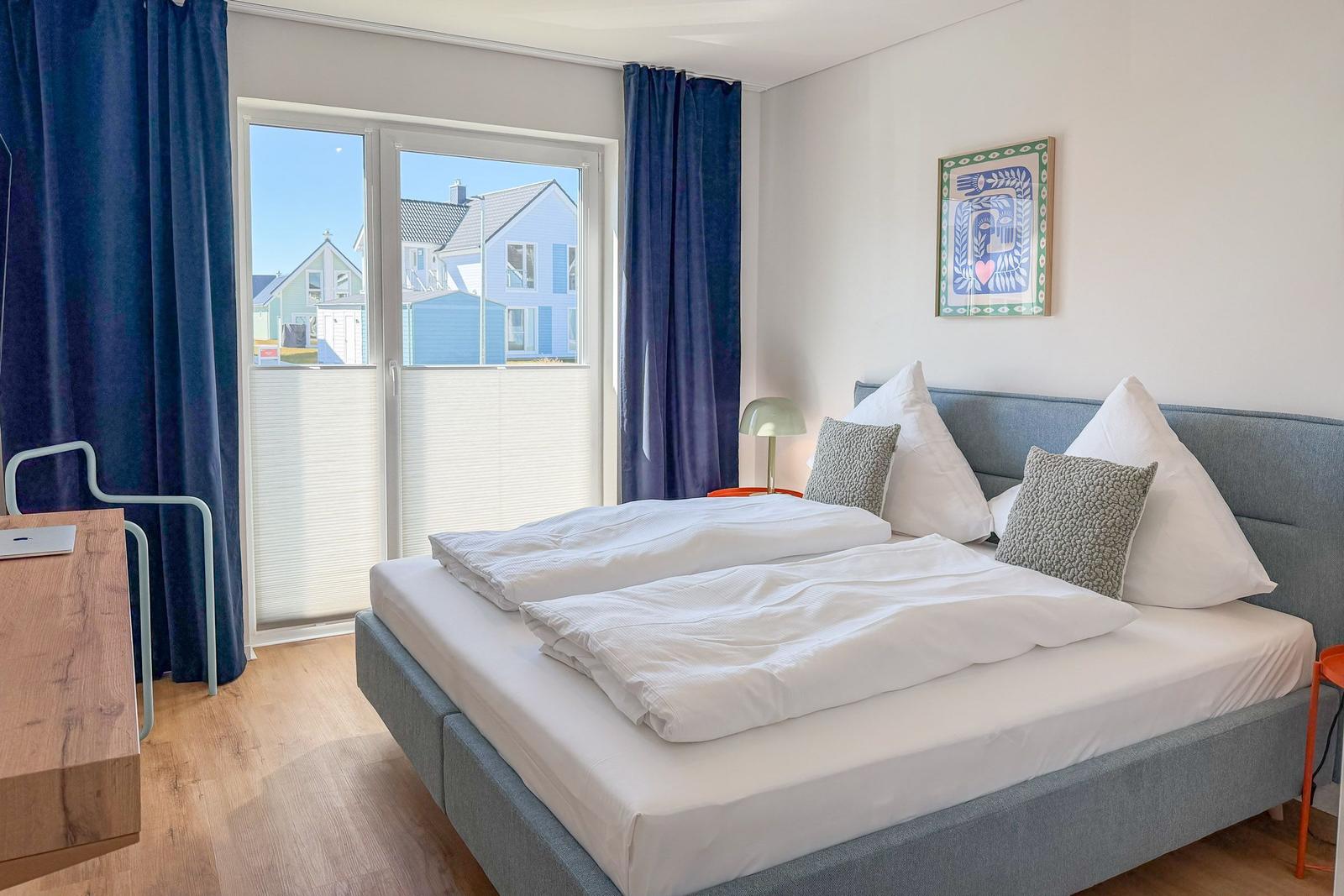 Doppelbett mit weißen Bettwäsche, blauen Vorhängen und Blick auf Häuser.