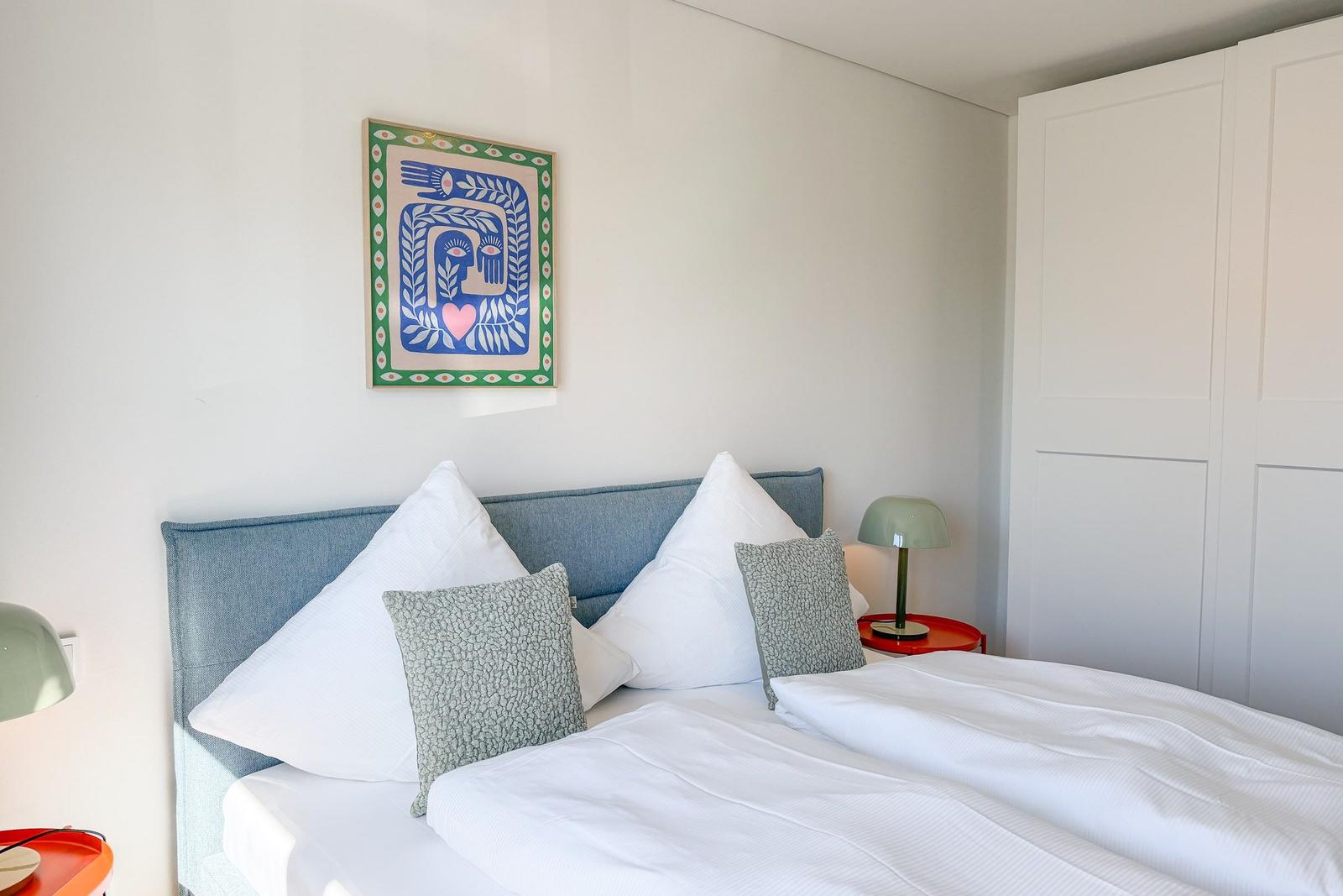 Schlafzimmer mit Bett, Kissen, Nachttisch und Wandbild.