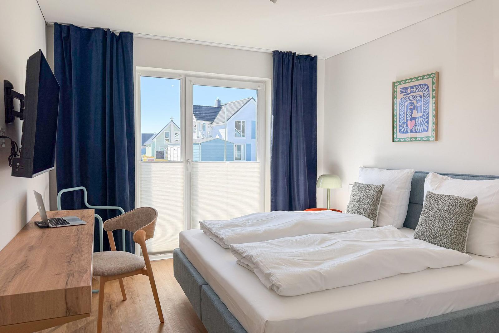 Schlafzimmer mit Bett, Schreibtisch, TV und Fensterblick auf Häuser.