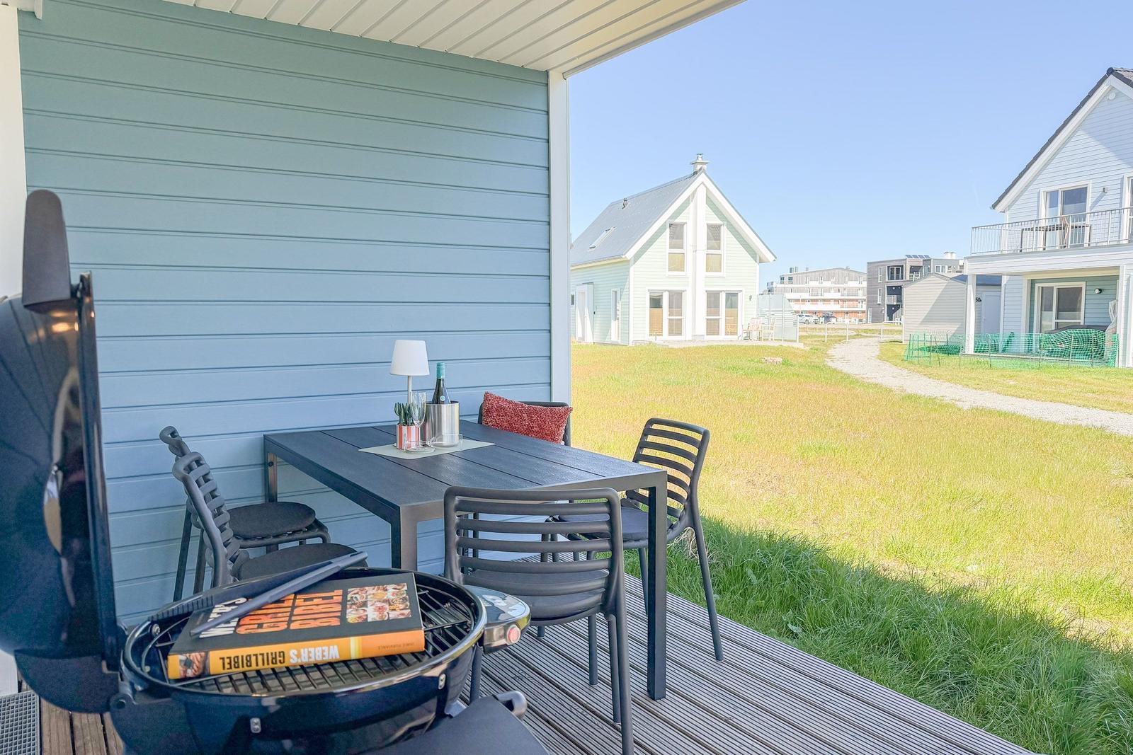 Terrasse mit Tisch, Stühlen und Grill. Blick auf Grasfläche und Häuser.