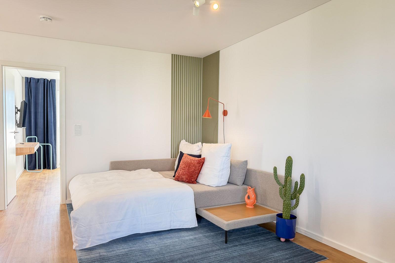 Schlafzimmer mit Bett, Sofa, Kaktus und Holzboden. Tür führt in einen angrenzenden Raum.