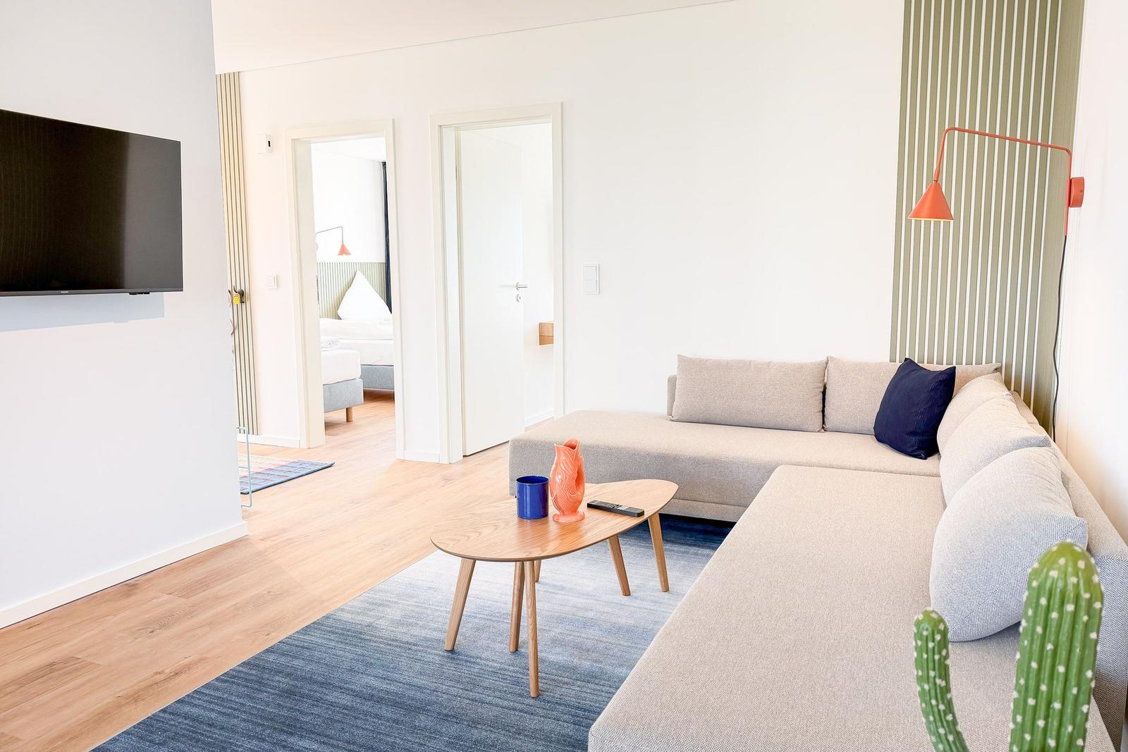Wohnzimmer mit Sofa, Couchtisch und Fernseher. Blick in Schlafzimmer und Bad.