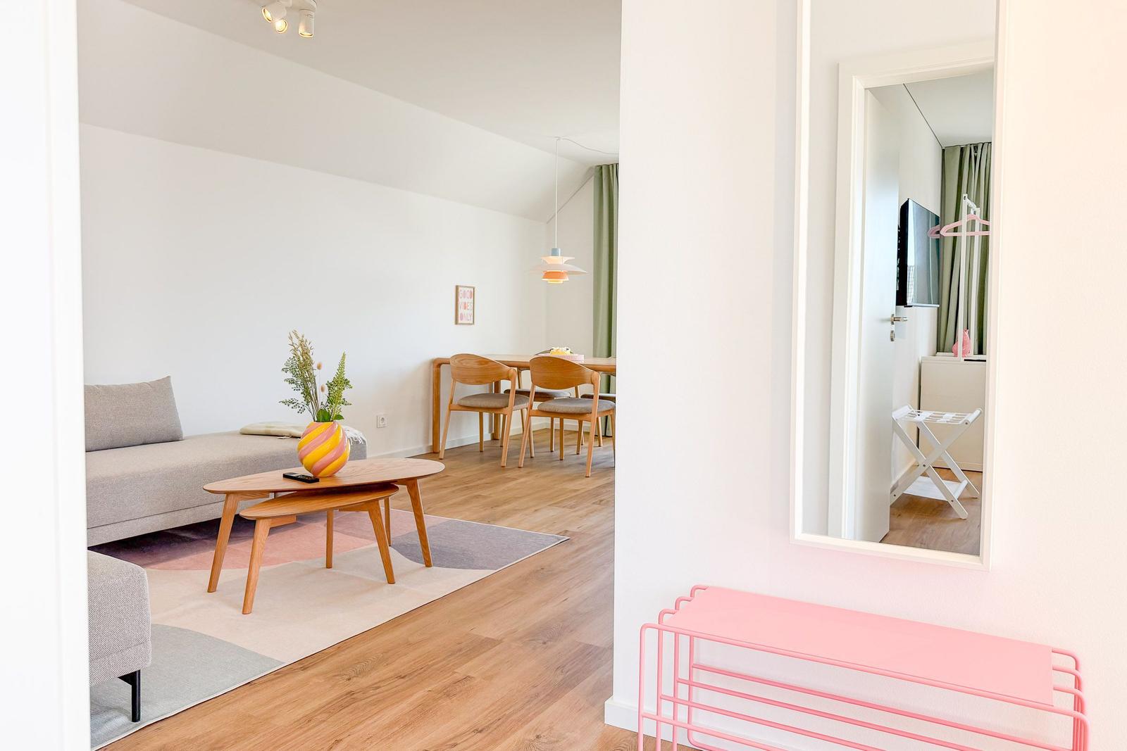 Wohnbereich mit Sofa, Esstisch und Spiegel. Pinkes Garderobenregal vorne.