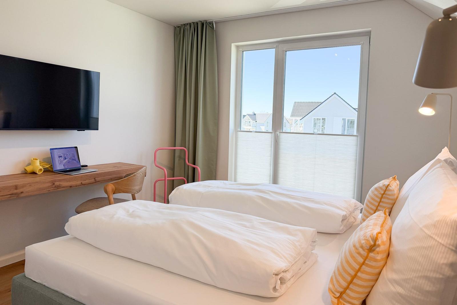 Zwei Betten, Schreibtisch mit Laptop, TV und Fenster mit Blick auf Häuser.