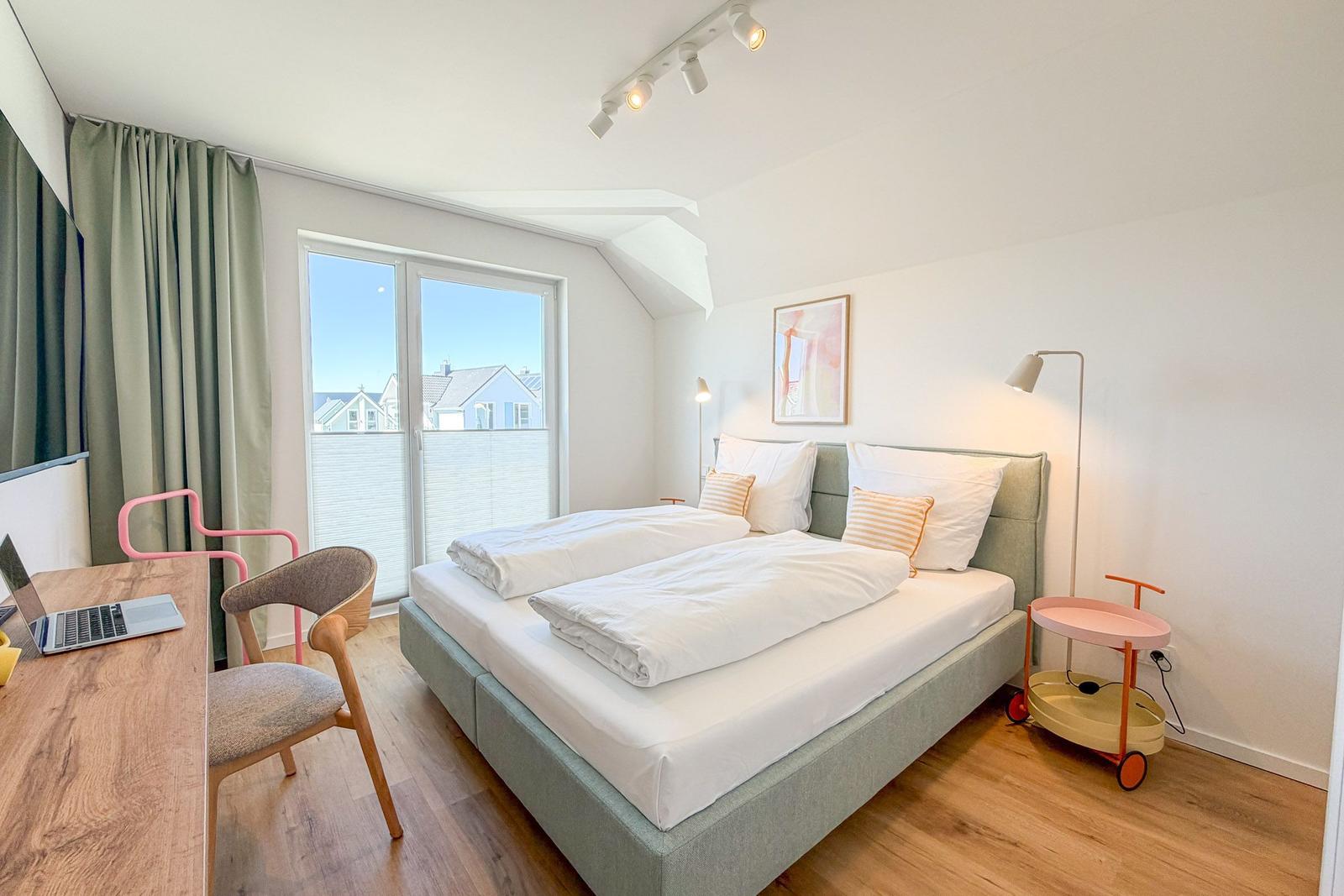 Schlafzimmer mit Doppelbett, Schreibtisch und Fensterblick auf Wohngebiet.