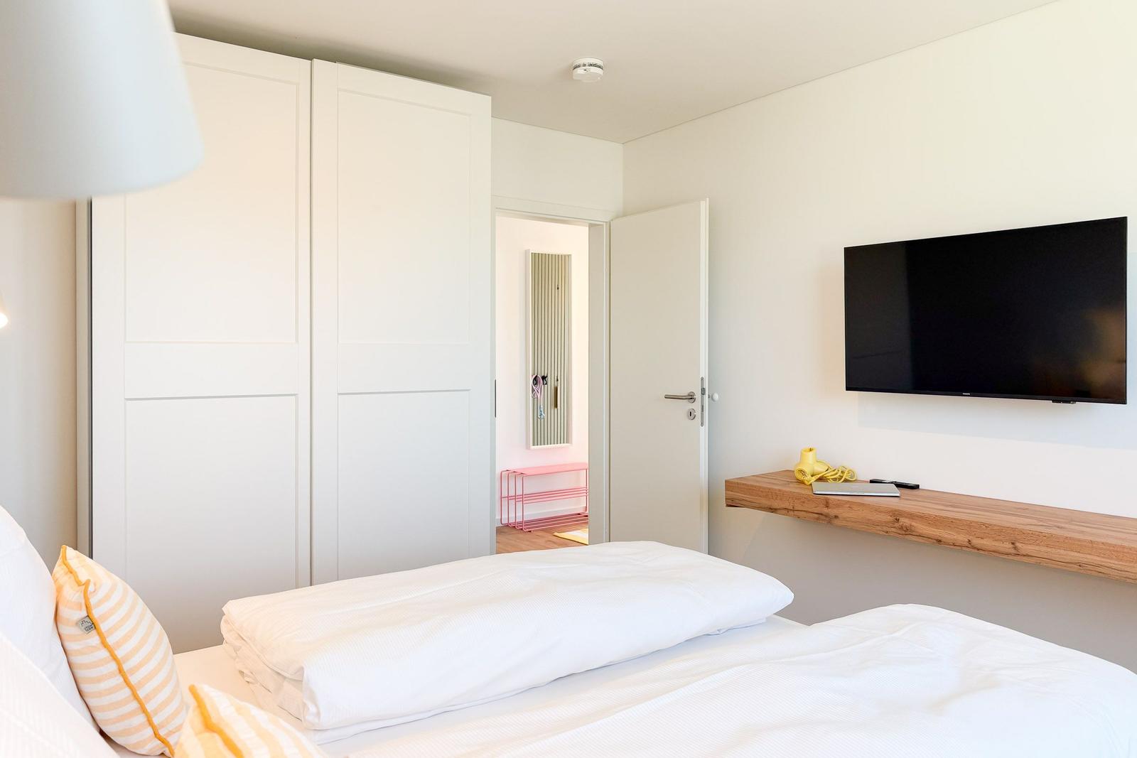Schlafzimmer mit Bett, Kleiderschrank und Wand-TV. Tür führt in einen Flur.