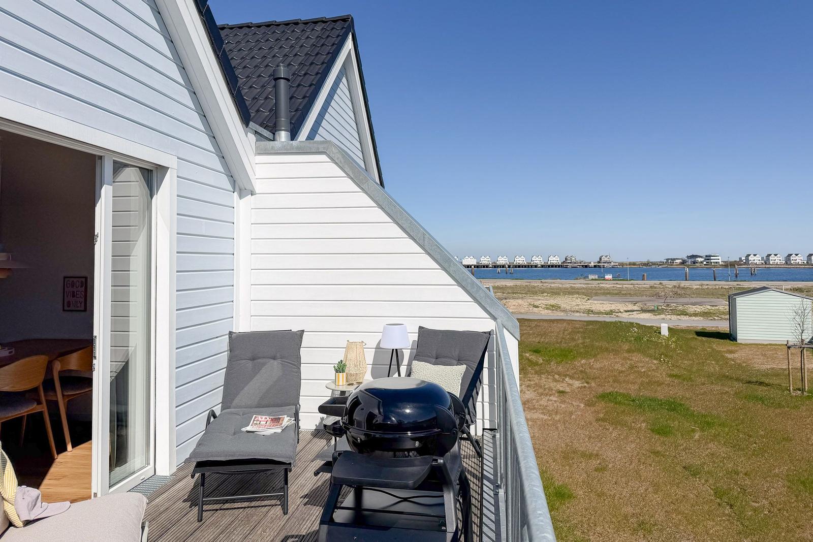 Terrasse mit Liegestühlen und Grill, Blick auf Wasser und Strand.