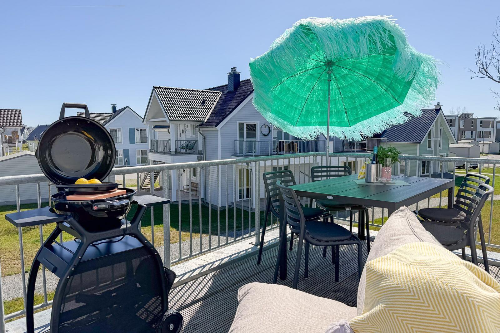 Terrasse mit Grill, Tisch, Stühlen und grünem Sonnenschirm. Hintergrund: Wohnhäuser.