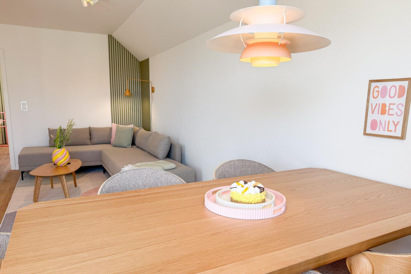 Esszimmer mit Holztisch und Sofa im Hintergrund. Deckenleuchte und Wandbild sichtbar.