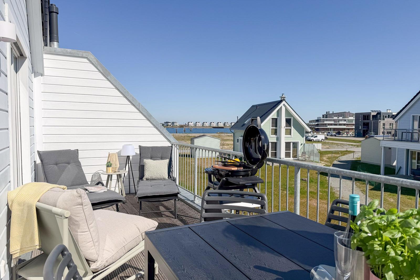Terrasse mit Sitzgelegenheiten, Grill und Tisch. Blick auf Wasser und Häuser.