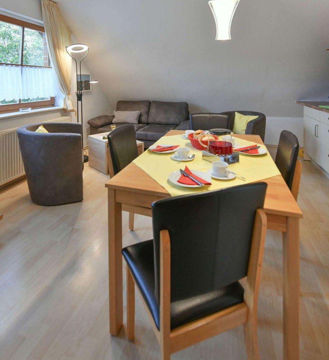 Esszimmer mit Tisch, Stühlen und Sofa im Hintergrund. Fenster mit Vorhängen.