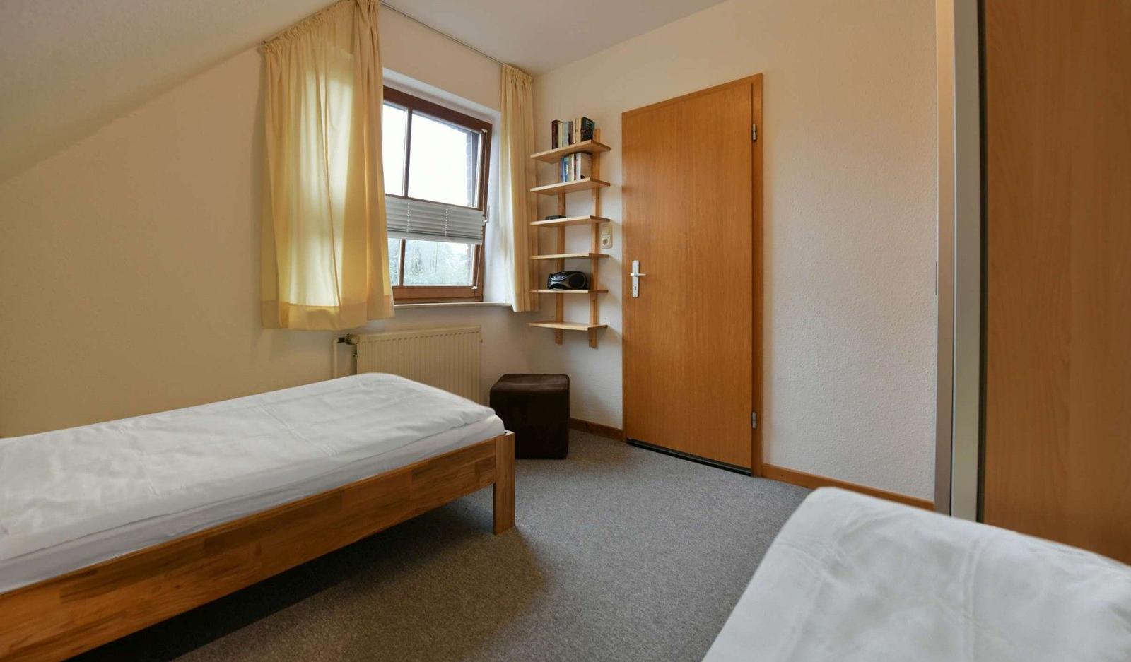 Zimmer mit zwei Betten, Fenster, Bücherregal und Tür.