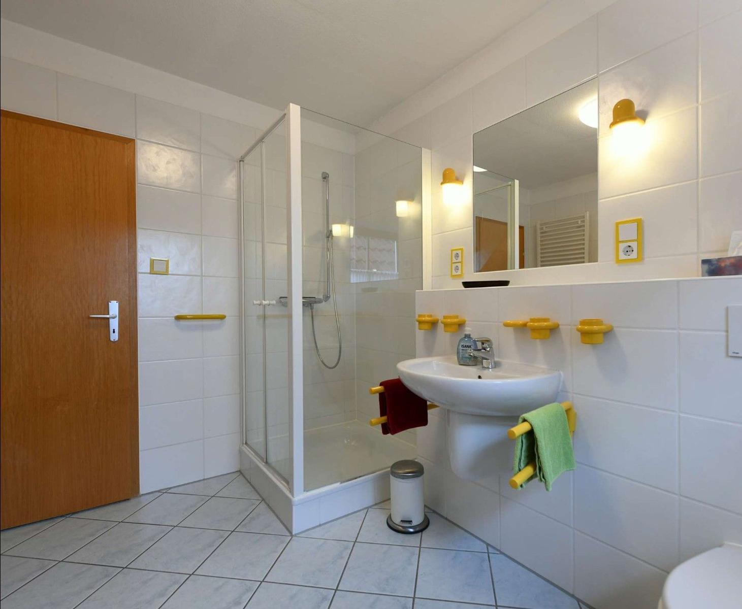 Badezimmer mit Dusche, Waschbecken und Spiegel. Gelbe Halterungen und Handtuchhalter.