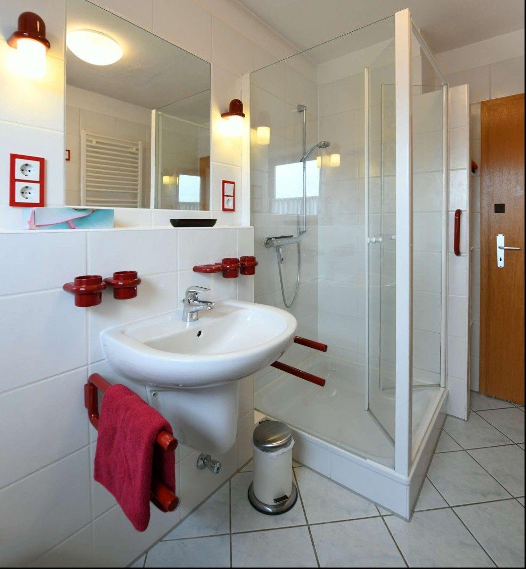Badezimmer mit Dusche, Waschbecken und Spiegel. Rote Haltegriffe und Handtuchhalter.