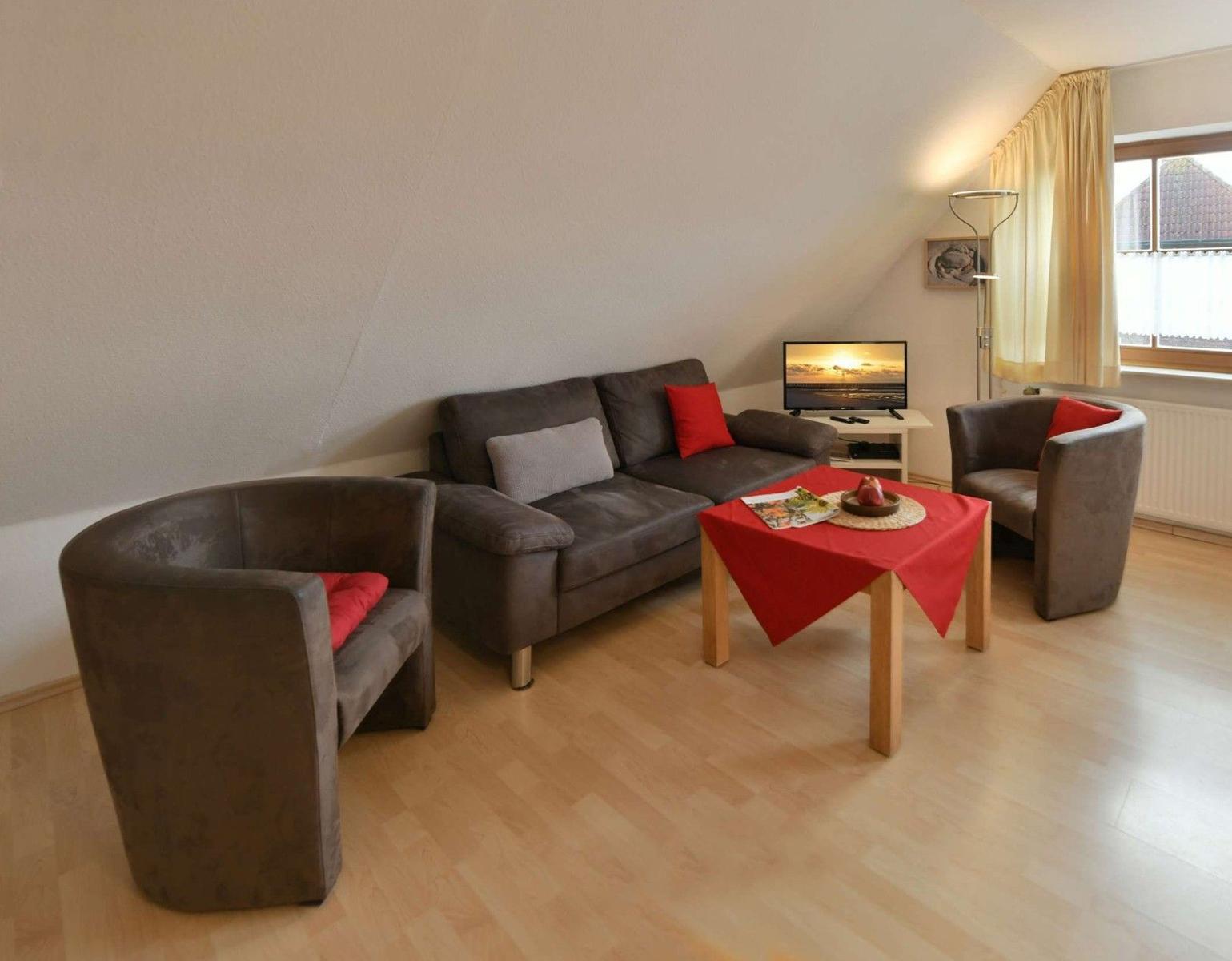 Wohnzimmer mit Sofa, zwei Sesseln, Tisch und Fernseher. Holzboden und Fenster mit Vorhängen.
