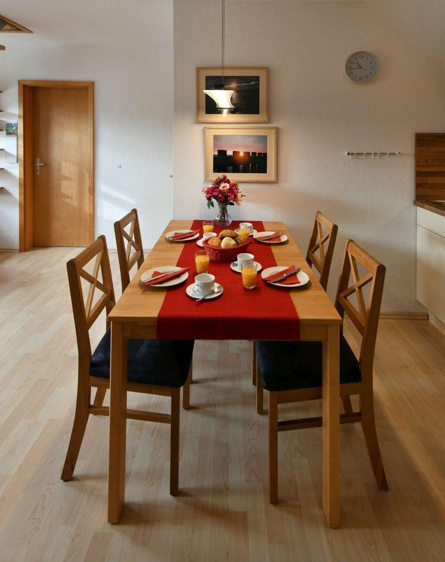 Esszimmer mit Holztisch, sechs Stühlen und roter Tischdecke. Wandbilder und Uhr sind sichtbar.
