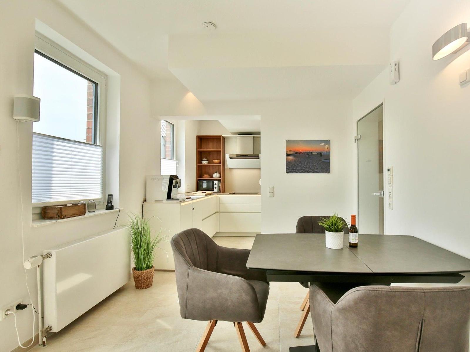Essbereich mit Tisch und Stühlen, Küchenbereich im Hintergrund, Fenster mit Jalousie.