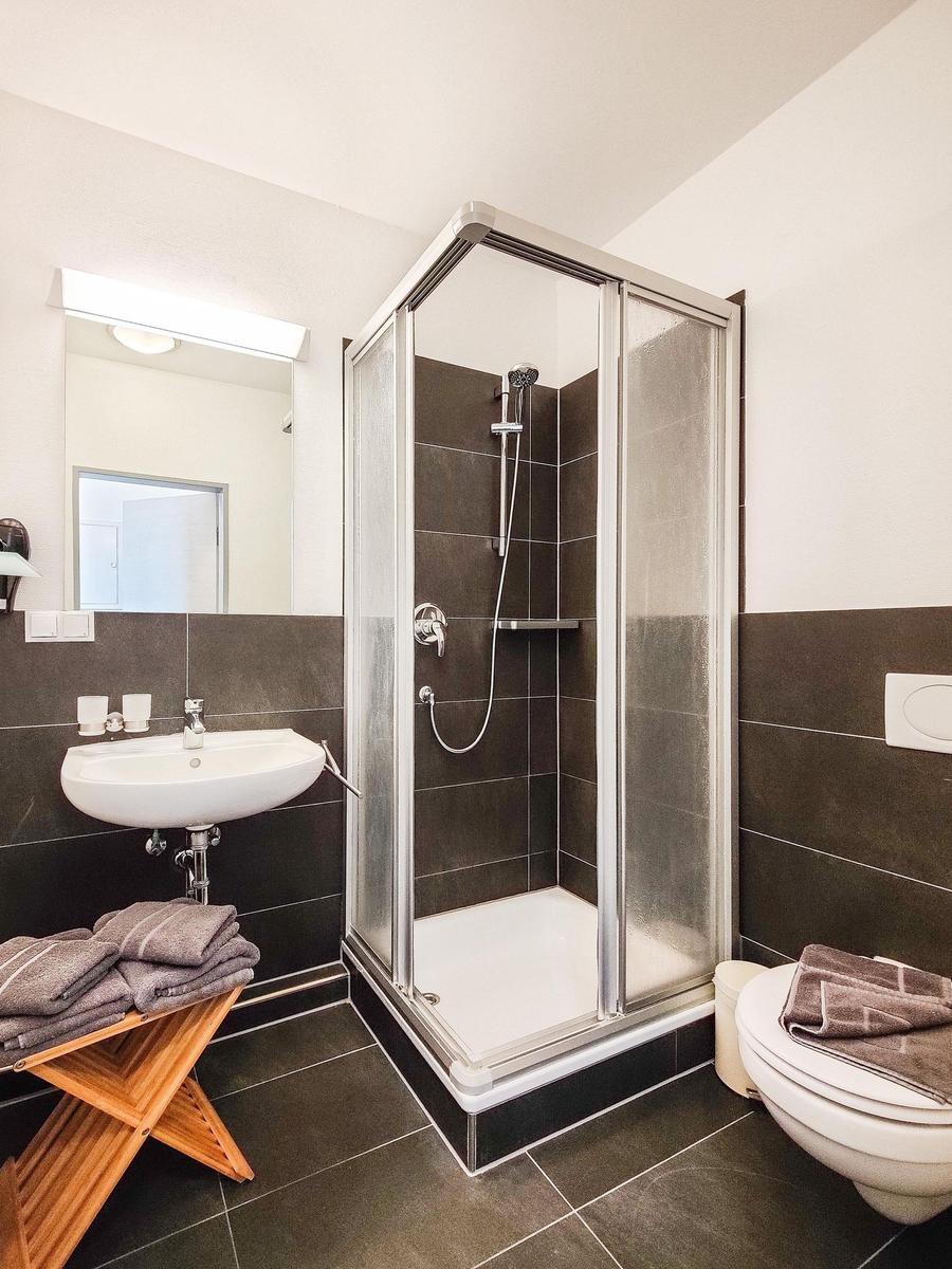 Badezimmer mit Dusche, Waschbecken und Toilette. Fliesen und Holzstuhl sichtbar.