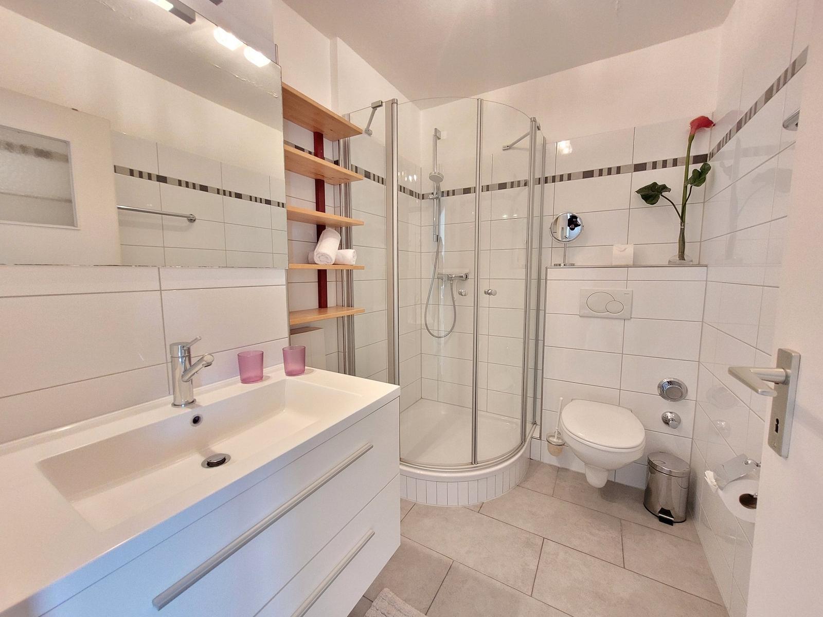 Badezimmer mit Dusche, Waschbecken und Toilette. Weiße Fliesen und Holzregale.