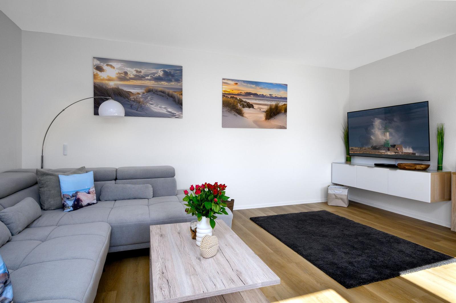 Wohnzimmer mit grauem Sofa, Holztisch und Wandbildern. Fernseher auf weißem Schrank.