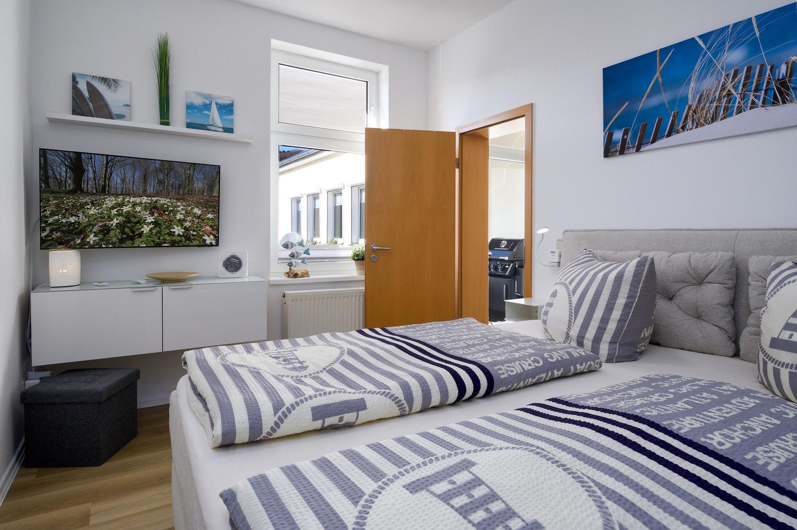 Schlafzimmer mit Bett, Fernseher und Fenster. Wandbild und Kissen mit Segelbootmotiv.