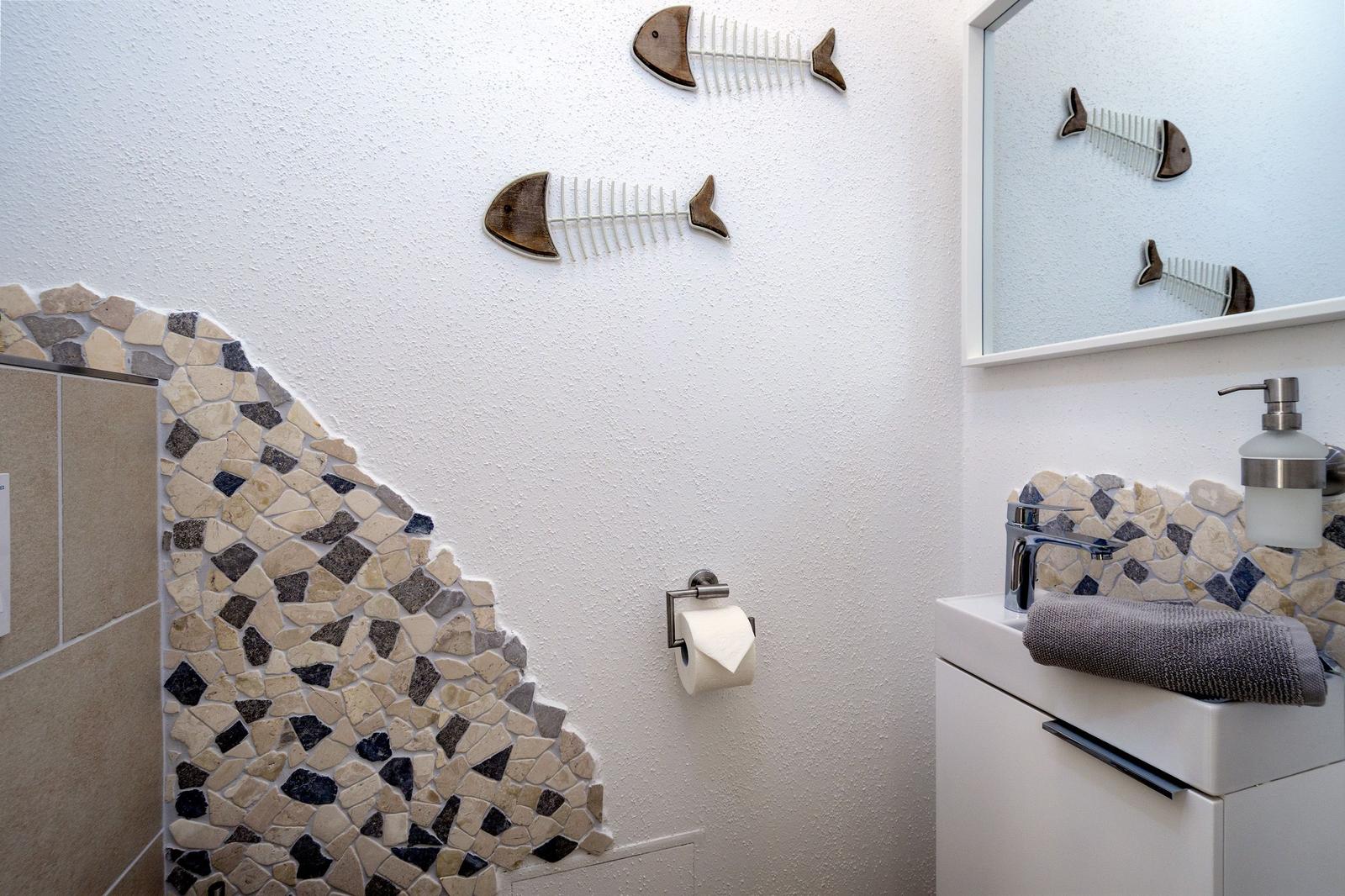 Badezimmer mit Mosaikwand, Toilettenpapierhalter und Spiegel mit Fischdekoration.