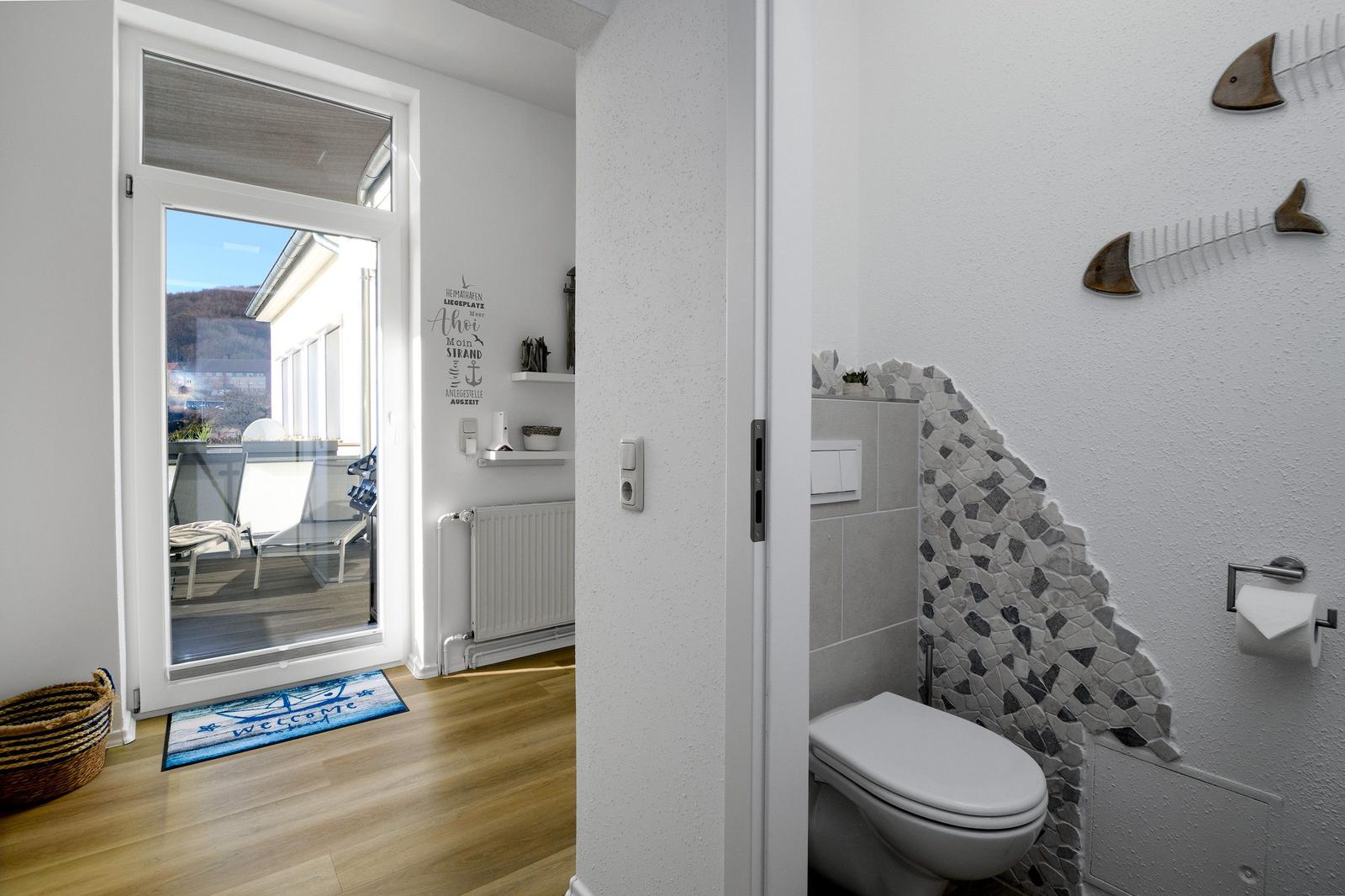 Badezimmer mit Toilette und Mosaikwand. Durchgang zur Terrasse mit Sitzgelegenheiten.