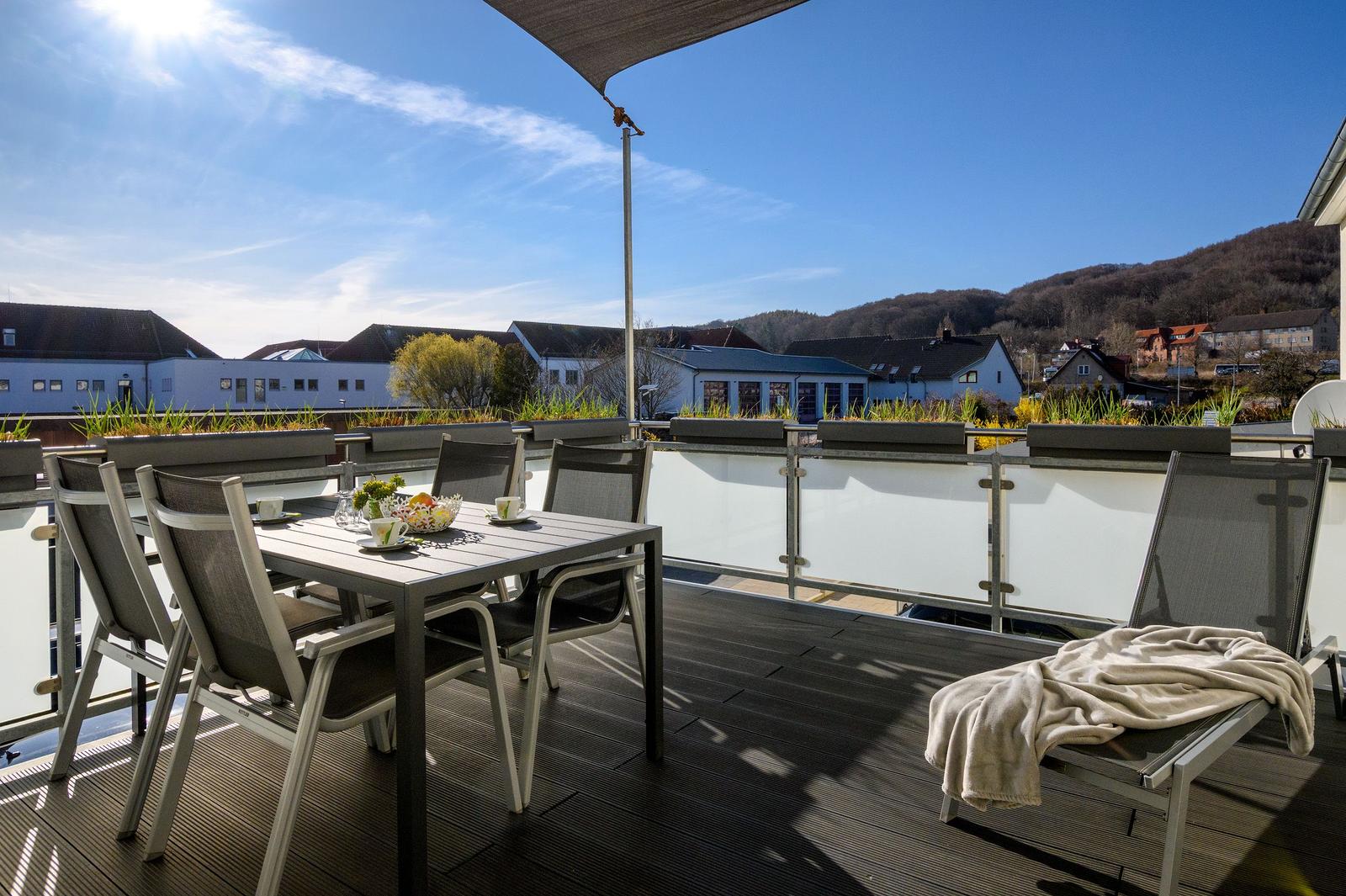 Terrasse mit Tisch, Stühlen und Liege. Blick auf Häuser und Hügel unter blauem Himmel.