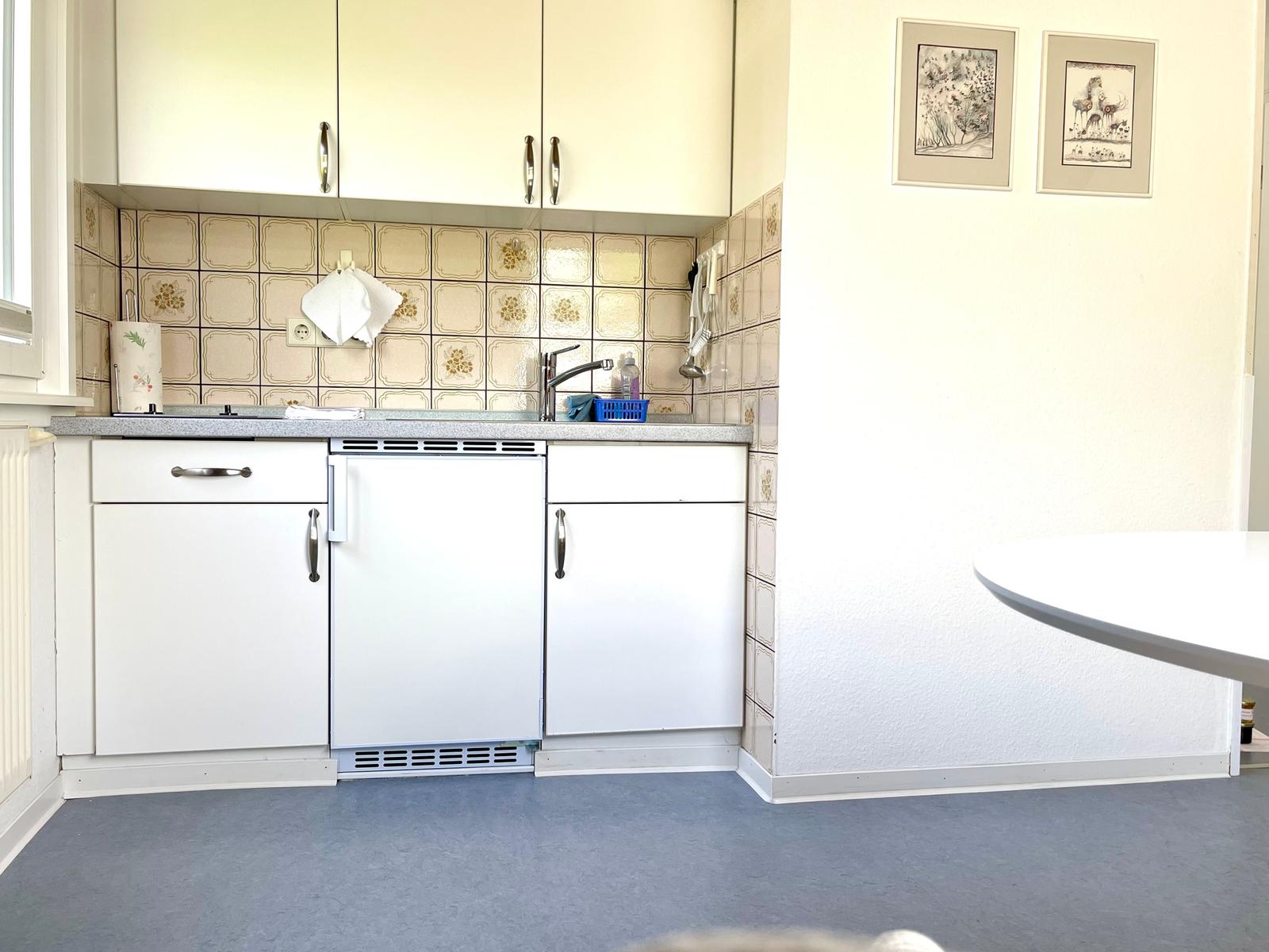 Küche mit weißen Schränken, Fliesenwand und Arbeitsplatte. An der Wand hängen zwei Bilder.