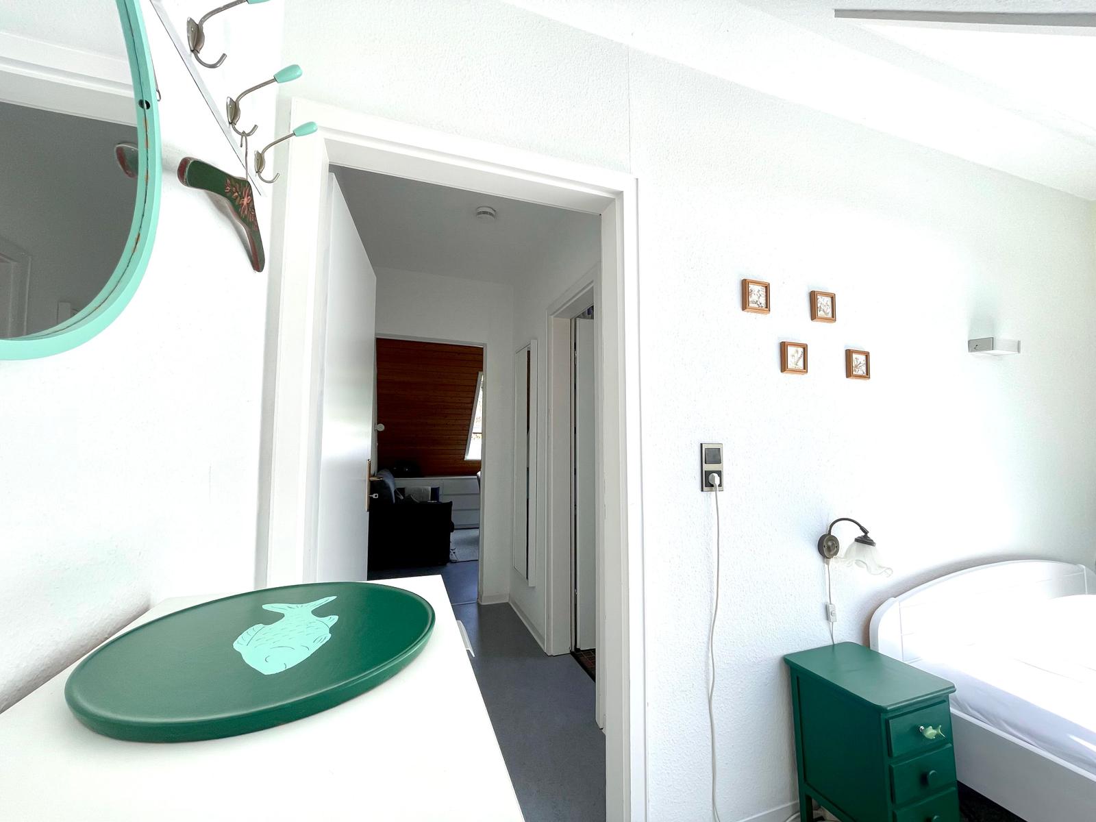 Schlafzimmer mit grünem Nachttisch und Spiegel. Durchgang zu Wohnbereich mit Holzwand.