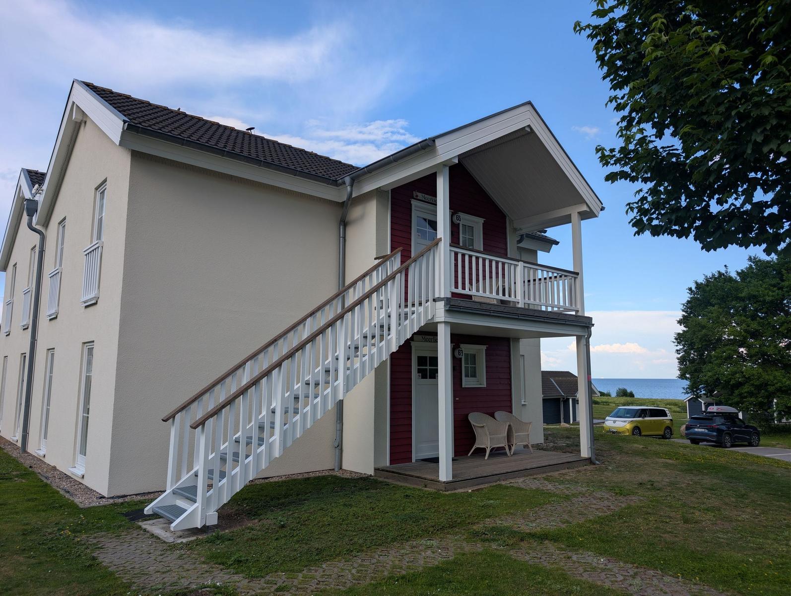Zweistöckiges Haus mit Balkon und Außentreppe. Blick aufs Meer im Hintergrund.