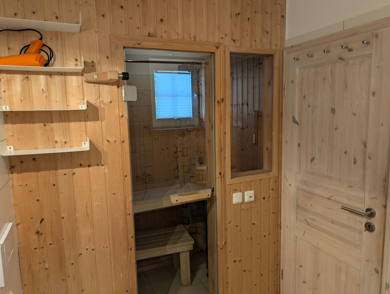 Sauna mit Holzwand, Sitzbank und Fenster. Orange Haartrockner auf Regal.