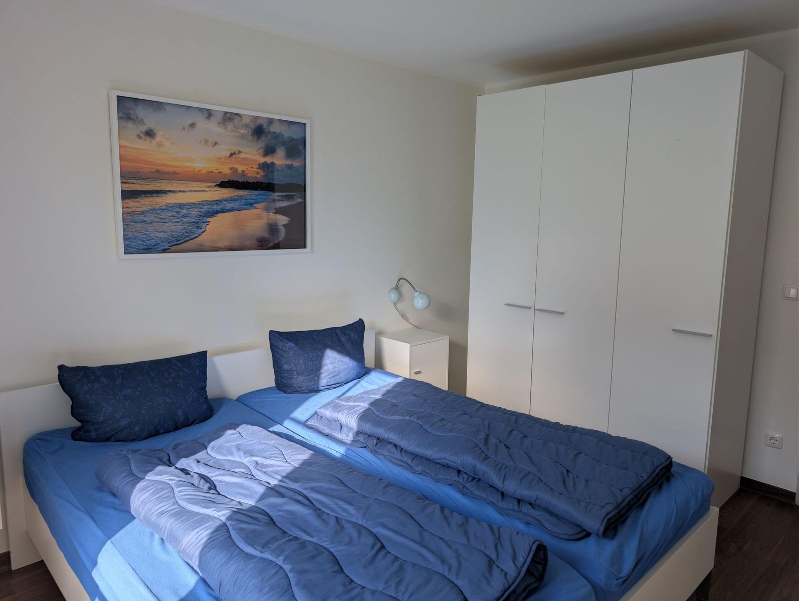 Zimmer mit Doppelbett, weißen Schränken und Wandbild mit Strandmotiv.