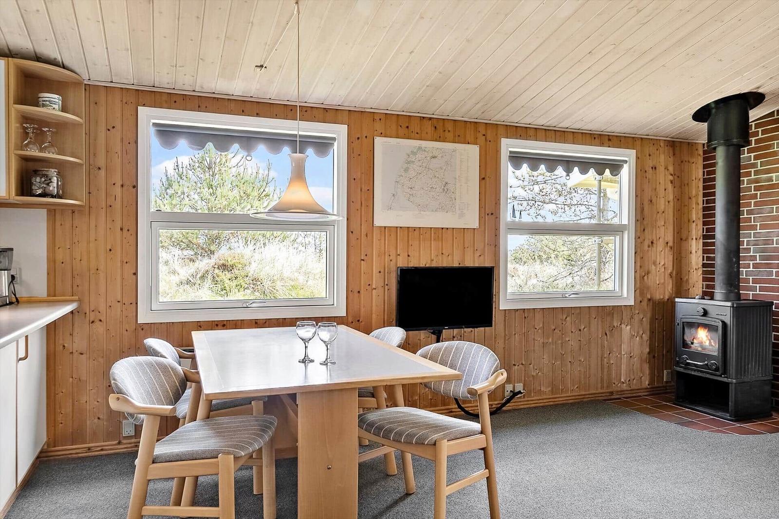 Esszimmer mit Holzwand, Tisch, Stühlen, Kaminofen und Fenstern mit Blick auf Außengelände.