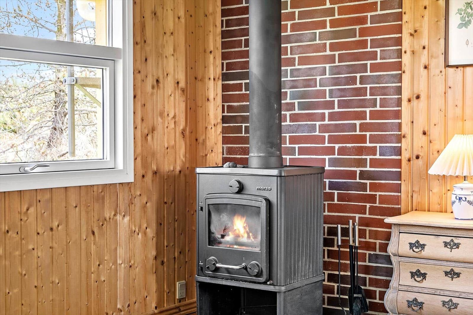 Kaminofen mit brennendem Feuer, Holzwand und Backsteinwand, Fenster mit Blick nach draußen.