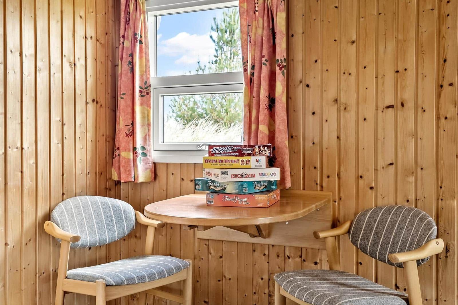 Zwei Stühle um einen runden Tisch mit Brettspielen. Holzwände und Fenster mit Vorhängen.