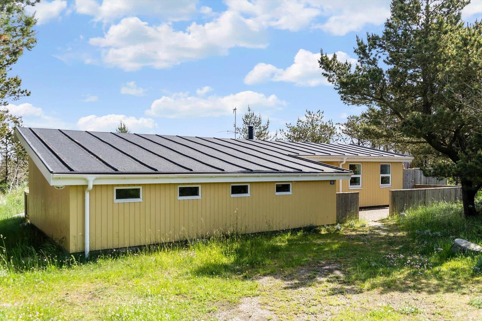 Ein gelbes Ferienhaus mit dunklem Dach steht in einer grünen Umgebung unter blauem Himmel.