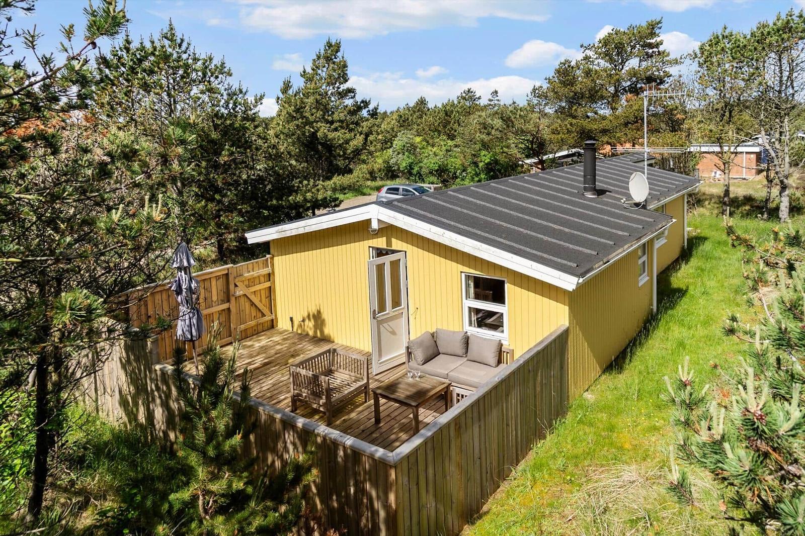 Gelbes Ferienhaus mit Terrasse, Garten und Grillplatz. Umgeben von Bäumen und Gras.