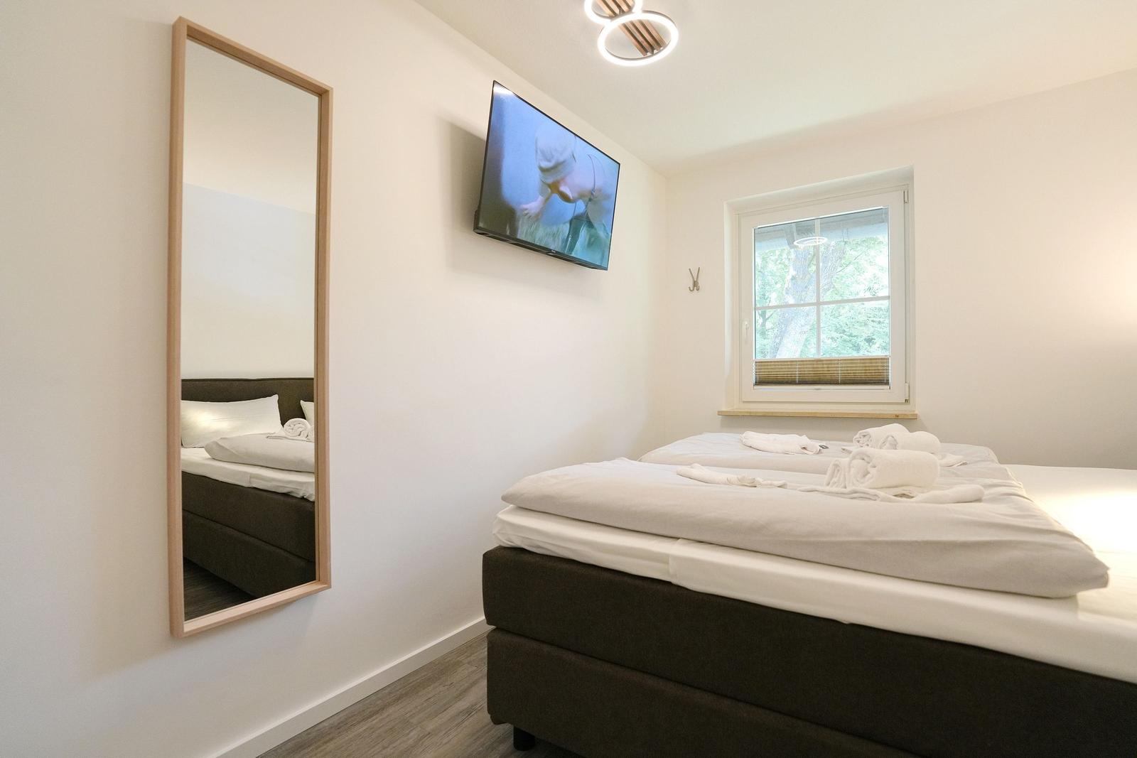 Schlafzimmer mit Bett, Spiegel und Wand-TV. Fenster mit Blick auf Bäume.