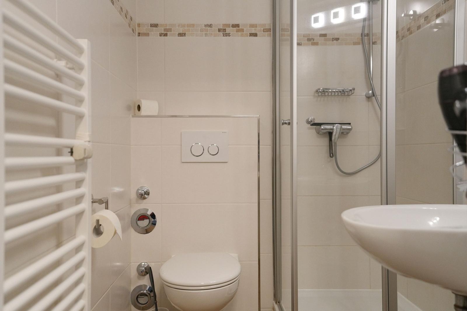 Badezimmer mit Dusche, Toilette und Waschbecken. Weiße Fliesen und Handtuchheizung.