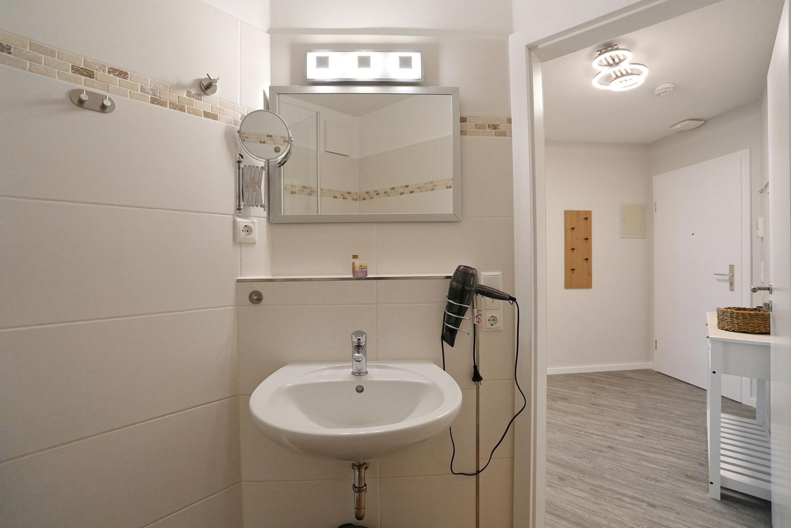 Badezimmer mit Waschbecken, Spiegel und Haartrockner. Blick in angrenzenden Raum.