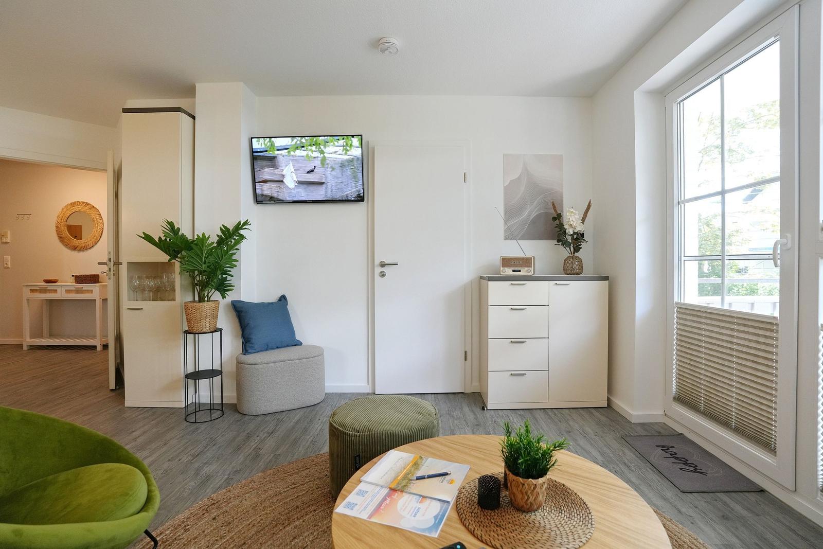 Wohnzimmer mit TV, Pflanzen und Sitzgelegenheiten. Große Fensterfront mit Blick nach draußen.