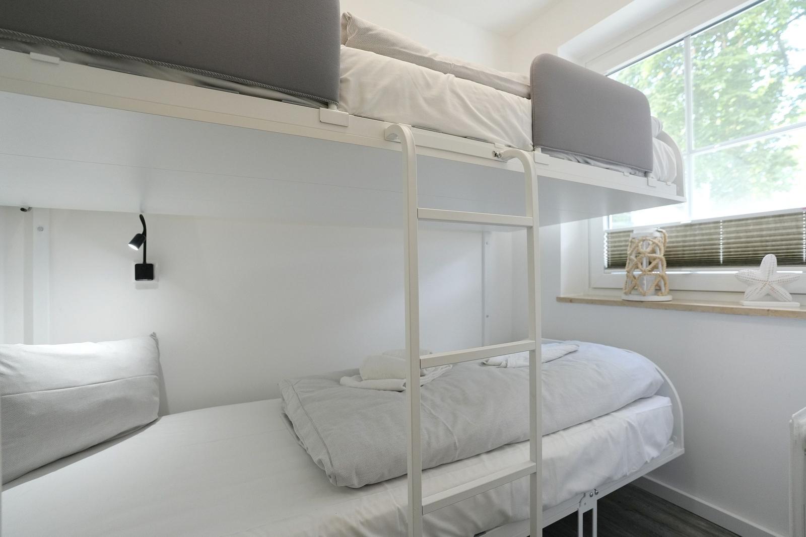 Zwei Etagenbetten mit grauen Matratzen und weißen Laken. Fenster mit Jalousie und Dekoration.