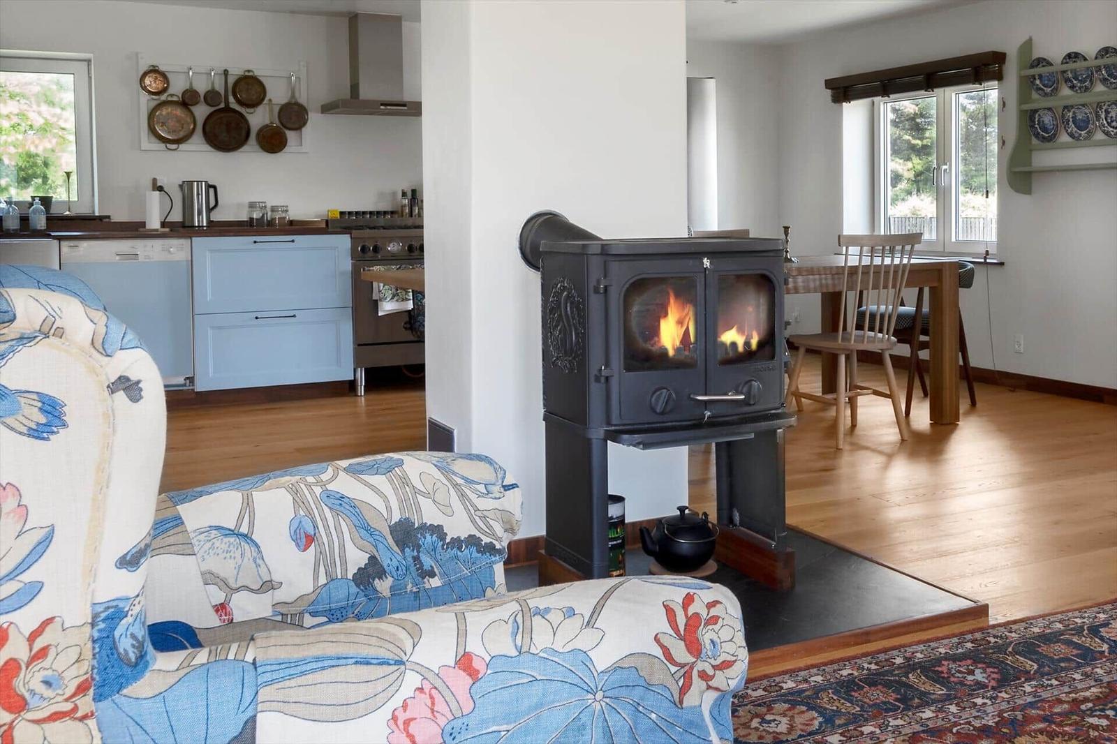 Wohnzimmer mit Kaminofen, Küchenbereich und Essbereich mit Holzboden und Fenstern.