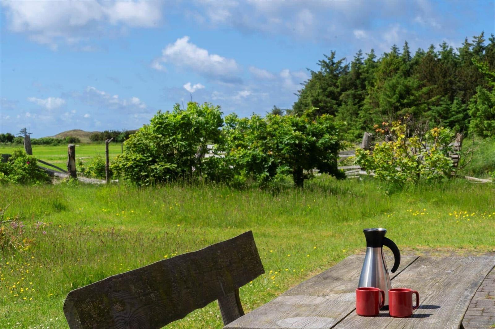 Holztisch mit Thermoflasche und zwei Tassen auf Terrasse. Grüner Garten und Wald im Hintergrund.