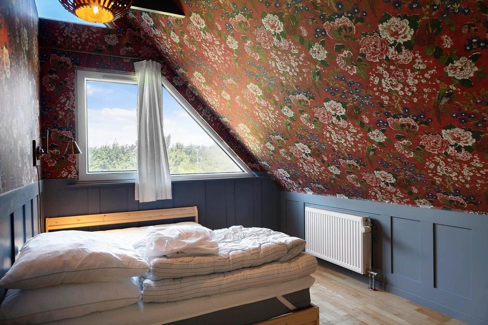 Schlafzimmer mit floraler Tapete, Holzbett und Dachfenster mit Blick auf Wald.