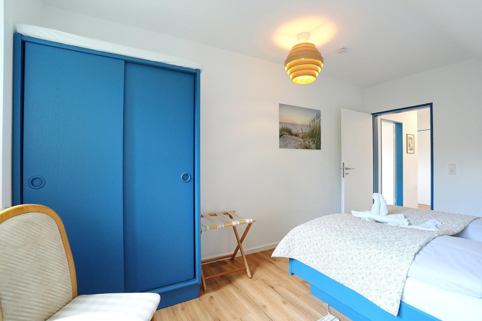 Schlafzimmer mit blauem Schrank, Bett und Holzboden. Deckenleuchte und Wandbild sind sichtbar.