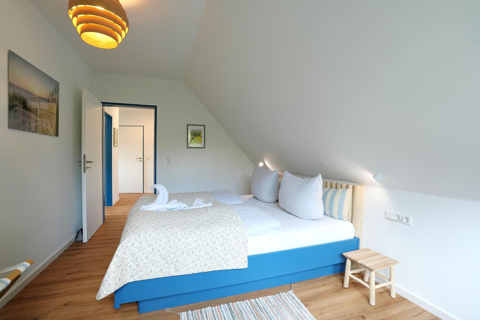 Schlafzimmer mit blauem Bett, Holzboden und schräger Decke. An der Wand hängt ein Bild.